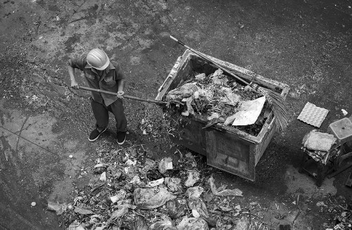 A worker wearing a hard hat and dark work clothes is using a long-handled tool to clear rubbish from the ground next to a large, open metal skip filled with mixed waste materials. The skip is positioned on a paved surface, with scattered debris and trash bags visible nearby. The waste inside the skip appears to include paper, plastic, and other general household or commercial refuse. The surrounding environment seems to be an outdoor area, possibly part of a street or industrial space, with a rough textured ground and some additional objects or materials placed on the right side of the image. The scene is captured in black and white, emphasizing the textures of the waste and surface, conveying a professional scene of rubbish removal that could be associated with services offered by Office Clearance Hackney or similar rubbish removal companies in the Hackney area.