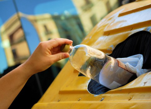 A person’s hand is shown holding a clear plastic water bottle above a yellow vehicle, possibly a van or truck used for rubbish collection, with the bottle appearing ready to be placed into a cargo area. The vehicle is situated outdoors, with part of a cityscape visible in the background, including building facades and windows, suggestive of an urban environment in Hackney or a similar London borough. The vehicle’s surface has a smooth, glossy finish, and the cityscape reflects subtly in the background. The image emphasizes the act of waste disposal or collection, consistent with professional rubbish removal services, such as those offered by Office Clearance Hackney. The scene captures the moment of emptying or loading waste or recyclable items, with the vehicle positioned on a paved or tarmac surface. The overall lighting is natural, indicating daytime, and the environment suggests an active, urban setting focused on waste management or rubbish clearance activities in the Hackney area.