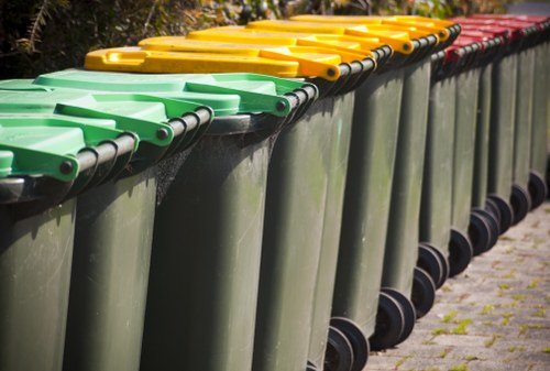 A row of large, rectangular waste bins with ergonomic, hinged lids—green in the front, followed by yellow and red lids—placed on a pavement in an outdoor setting, possibly in Hackney. The bins are made of durable plastic with a smooth surface finish and are positioned side by side, aligned along the curb or pavement edge. The background features some greenery and trees, suggesting an urban or suburban environment. The scene appears to be during daylight, with natural light illuminating the bins and the surrounding area. This setup is typical for waste disposal and rubbish collection services in the Hackney area, operated by a company like Office Clearance Hackney, offering reliable rubbish removal solutions for local businesses and residents.