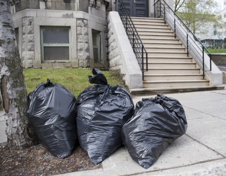 Three large black rubbish bags made of plastic and tightly tied are placed on the pavement near the base of a tree in an urban environment, with a concrete staircase and metal handrails leading up to a building entrance visible in the background. The bags appear to contain waste material related to office clearance or domestic rubbish removal, set against the backdrop of a city street possibly in Hackney, London, as suggested by the location context. The scene is outdoors during daylight with natural lighting, and the bags are positioned on a sidewalk adjacent to a small patch of grass and the building's exterior wall. The presence of these filled rubbish bags indicates the ongoing process of waste collection or clearance typical of services provided by Office Clearance Hackney, with the bags prepared for disposal or removal from the premises.