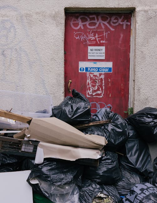 A red, weathered metal dumpster with signs of rust and peeling paint is positioned in a narrow alleyway corner, adjacent to a dark green wall on the left and a textured concrete wall on the right. In 