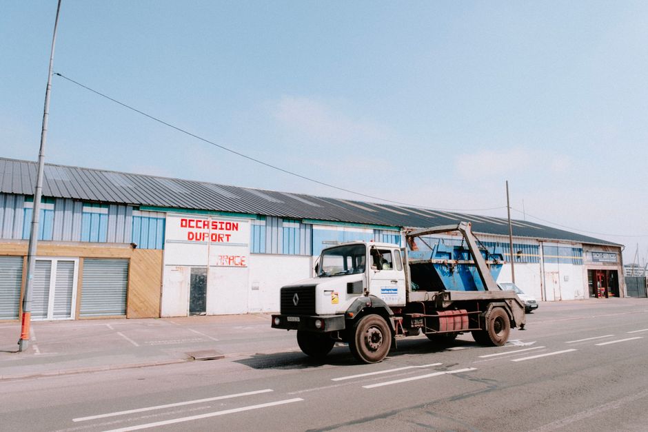 A small blue and yellow dump truck is parked on a paved street, with its bed tilted to unload a load of dark asphalt or gravel. The truck’s bed contains some brown fabric or clothing items laid on t