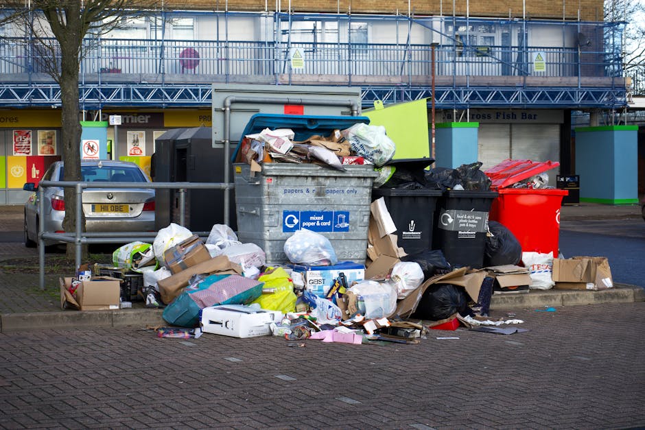 A large collection of mixed household waste, including black, black with white labels, and red rubbish bags, overflowing from open and closed wheelie bins placed on a paved sidewalk near a parking are
