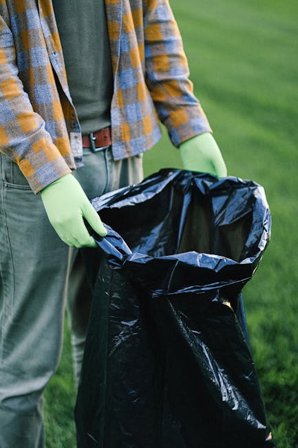 A large collection of mixed household waste, including black, black with white labels, and red rubbish bags, overflowing from open and closed wheelie bins placed on a paved sidewalk near a parking are