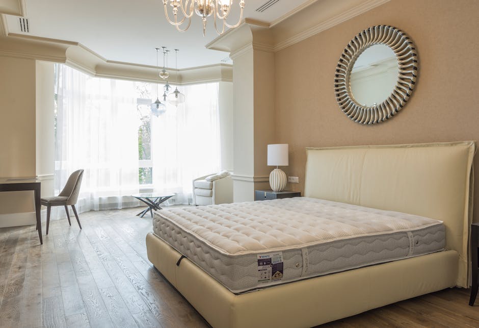 A bedroom interior featuring a large, plush off-white mattress resting on a matching bed frame with a padded headboard. The mattress has a textured surface with visible tufting and a manufacturer's la
