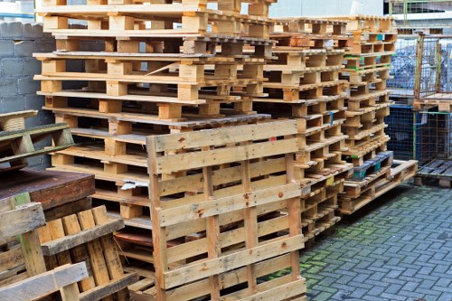 A large stack of untreated wooden pallets arranged vertically and horizontally on a paved surface, with some leaning against one another and others stacked neatly in a storage yard. The pallets are made of light, natural wood with visible grain texture and some darker marks, indicating previous use. In the background, there is a chain-link fence and a partial view of a building structure, suggesting a commercial or industrial environment typical for rubbish removal services in Hackney. To the left, smaller wooden crates or platforms are positioned beside the pallets, all situated outdoors in natural daylight. The scene reflects equipment used in waste management or clearance operations, aligning with the services offered by Office Clearance Hackney in the local area.